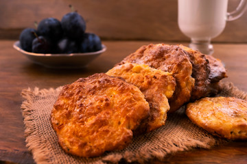 Cottage cheese cookies on a linen cloth napkin, milk in a glass cup, blue ripe plums in a porcelain plate, wooden background.