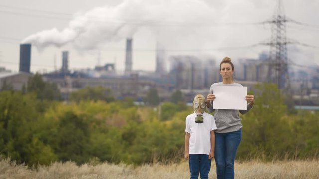 Mom And Son In A Gas Mask Stand In The Background Of A Factory And Smoking Pipes. In The Hands Holding A Sign