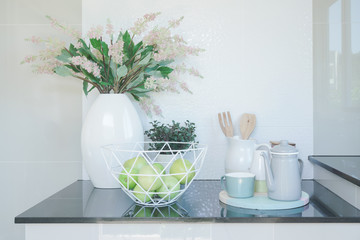 Green apple in a modern basket, ceramic tea set and beautiful flower vase on black top counter in modern style kitchen interior