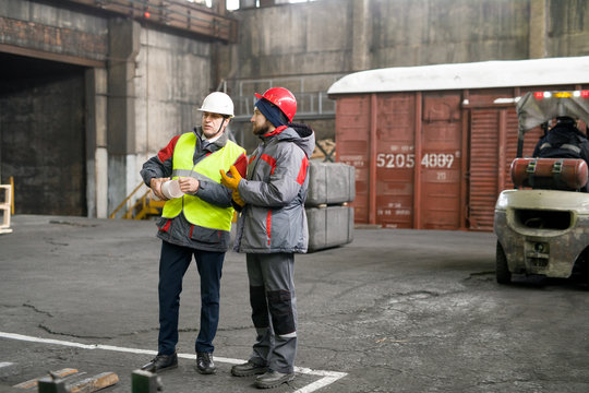 Full length portrait of two industrial workers wearing warm jackets and hardhats discussing production standing in workshop, copy space