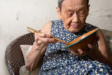90 up of healthy  old woman having lunch,front view..Portrait of a happy elderly woman in colorful dress  eating noodle with chopsticks by herself.