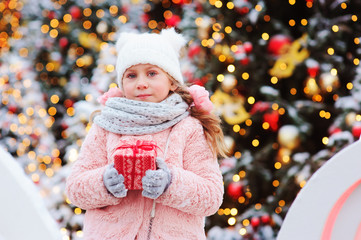 happy child girl holding christmas gift outdoor on the walk in snowy winter city decorated for new year holidays. Trees with christmas lights on background