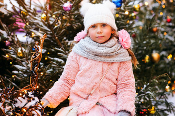 christmas portrait of happy child girl walking outdoor, snowy winter decorated trees on background. New Year Holidays in city.