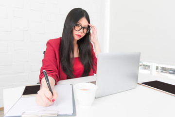 Asian businesswoman Wearing glasses and a red suit are sitting anxiously in front of the notebook in the modern office.