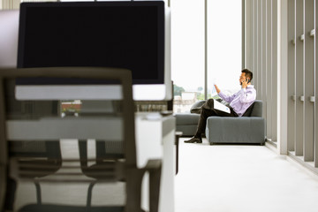 Young Asian businessman using smartphone on sofa there are documents in hand in the office where the computer is placed on the table in the modern office.