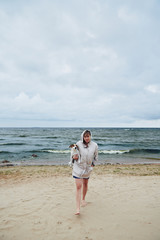 Beautiful young woman in warm jacket holding adorable dog and looking at camera while standing on sandy coast near stormy sea