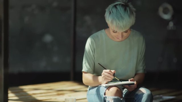An informal young artist in torn jeans, with piercings and short colored hair. She paints it with a brush, dabs it in the paint and does the smears. The studio has sunlight and beautiful shadows.