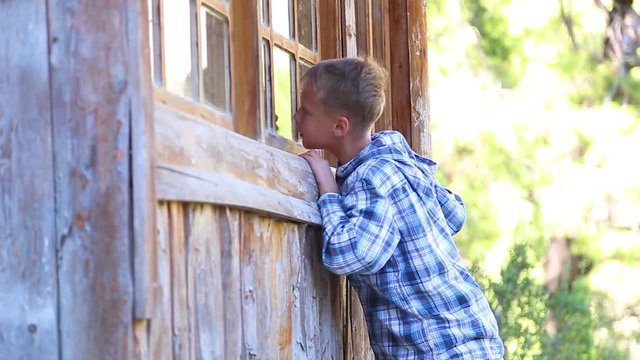 Lonely white kid trying to look inside of old abandoned wooden house through glass of window. Real time full hd video footage.