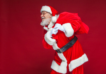 Christmas. Kind and tired Santa Claus in white gloves carries a red bag with gifts over his shoulder. Isolated on red background.