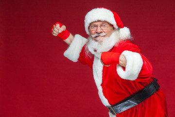 Christmas. Santa Claus with red bandages wound on his hands for boxing imitates kicks. Kickboxing, karate, boxing. Isolated on red background.