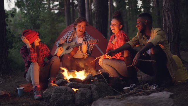 Cinemagraph loop - cheerful guy in casual clothing is playing the guitar while his male and female friends tourists are singing and smiling sitting around fire in forest.