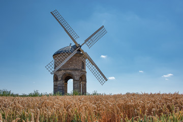 Fototapeta na wymiar A windmill stands in the summer sunshine. Located on top of a hill and surrounded by wheat. 