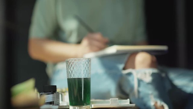 Glass with water, painted in green. Details of the artist's work. In the background, the silhouette of a woman artist in ragged jeans. She paints it, dabs the brush in a glass of water.