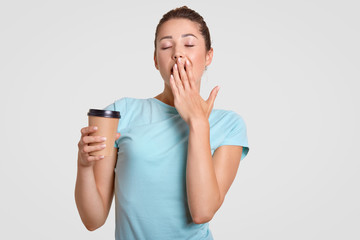 Sleepy woman yawns, covers mouth with hand, drinks hot coffee to feel refreshed, dressed in casual blue t shirt, isolated over white background. Female model with disposable takeaway coffee.