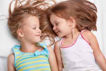 Two small sisters sleep together, look with positive expressions at each other, dressed in casual clothes, have good relationships. Best kid female friends have fun together in comfortable bed