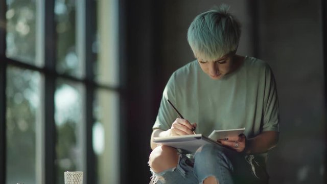 Young woman with a short haircut, colored hair, piercer holds on her knees an album for drawing. Artist paints with paintbrush, smiling in the process. Inspiration. An artist with a bright appearance.