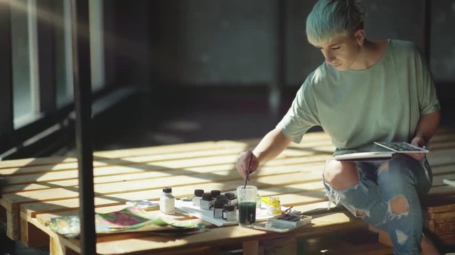 An informal young artist in torn jeans, with piercings and short colored hair. She paints it with a brush, dabs it in the paint and does the smears. The studio has sunlight and beautiful shadows.