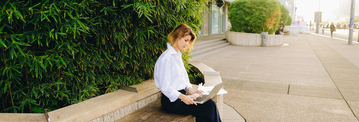 Female statistician working with color diagrams and laptop outdoors near green plant. Concept of working with theoretical or applied statistics. Young woman wears business clothes.