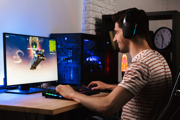 Portrait of young gamer man playing video games on computer, wearing headphones and using backlit...