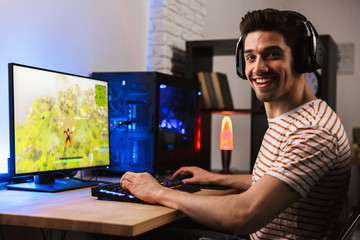 Portrait of happy smiling guy playing video games on computer, wearing headphones and using backlit colorful keyboard - obrazy, fototapety, plakaty