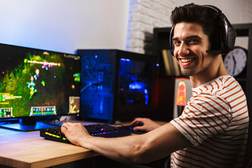 Image of cheerful gamer man playing video games on computer, wearing headphones and using backlit...