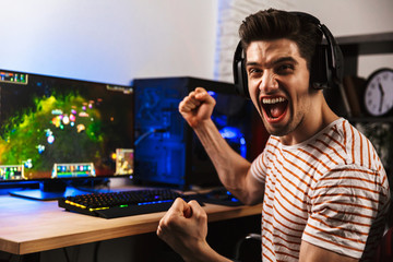 Portrait of happy man playing video games on computer, wearing headphones and using backlit...