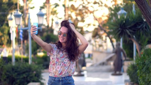 Brunette girl posing for selfie photo in tropical city. Cheerful woman in sunglasses taking selfie near palm trees. Female tourist taking mobile photo on resort. Joyful girl selfie
