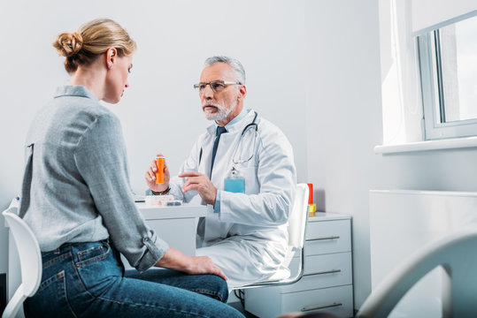 Serious Mature Male Doctor Pointing At Pills To Female Patient At Table In Office