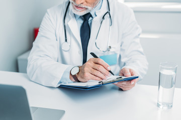 partial view of male doctor writing in clipboard and looking at laptop in office
