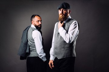 Two bearded men in strict suits standing one near one and looking on camera on black