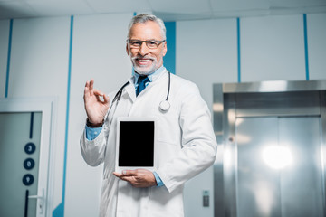 smiling mature male doctor with stethoscope over neck doing ok gesture and showing digital tablet with blank screen in hospital