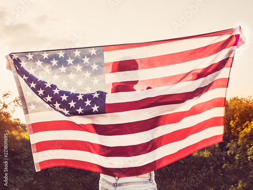 Attractive man holding a US flag against the background of the rays of the setting sun. View from the back. Preparing for the holidays
