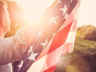 Attractive man holding a US flag against the background of the rays of the setting sun. View from...