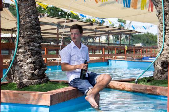 A young man in a white shirt is sitting near a palm tree in the pool with a bottle