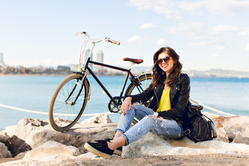 Cute brunette girl in sunglasses is sitting on stone near bike on sea background. She wears ripped jeans, black jacket, sunglasses, smiling to camera.