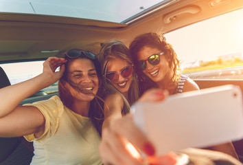 Three female friends enjoying traveling in the car. Sitting in rear seat and and making selfie. - obrazy, fototapety, plakaty