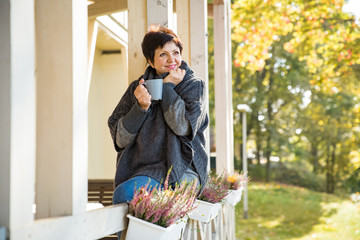 Mature attractive woman standing on Cozy wooden terrace with cup of hot coffee wrapped up in...
