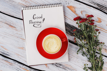 Good morning concept, flat lay. Cup of coffee, red flowers and notepad. White wood background, top view.