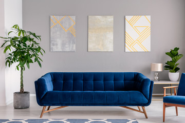 A dark blue velvet couch in front of a gray wall with graphic paintings in a modern living room...