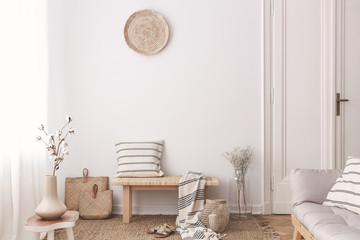 Flowers on wooden table near bench with pillow and blanket in white living room interior. Real photo