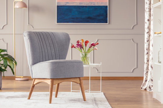 Grey chair on carpet next to table with flowers in elegant living room interior with lamp. Real photo