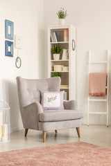 Cushion on grey armchair in bright living room interior with posters and pink carpet. Real photo