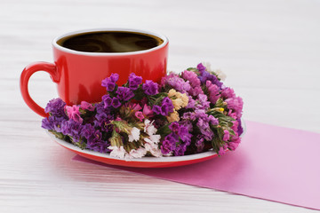 Red cup of hot coffee and statice limonium flowers. White wooden table surface. Close up.