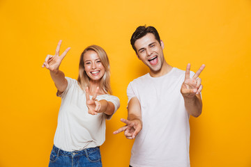 Excited young loving couple standing isolated over yellow wall background showing peace gesture.
