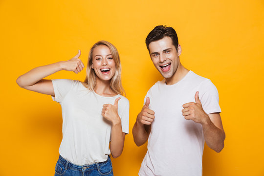 Young loving couple standing isolated over yellow wall background showing thumbs up gesture.