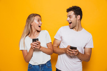 Happy excited young loving couple standing isolated over yellow wall background using mobile phones.