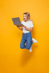 Excited young woman jumping isolated over yellow wall background using laptop computer.