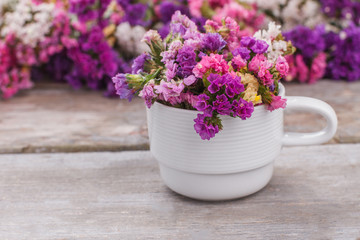 Multicolored statice flowers in white tea cup. Wooden desk background.