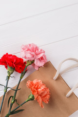 Three multicolored carnation flowers and shopping bag. White wooden surface background. Top view.