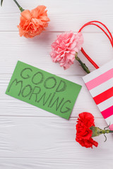 Colorful carnation flowers and good morning wish on note. Top view. White wooden background.